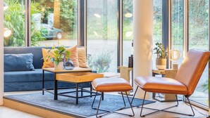 A modern living room with a sofa, a coffee table, and a leather chair. There is a large window with a view of greenery outside. - Best Western Bridge Hotel Lyon East (Jons)