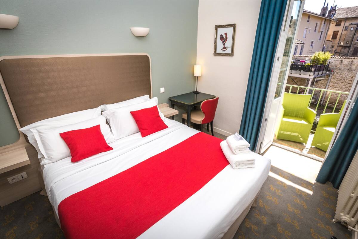 Ein Hotelzimmer mit einem großen Bett, einem kleinen Tisch, einem Stuhl und einem Balkon mit grünen Stühlen.
