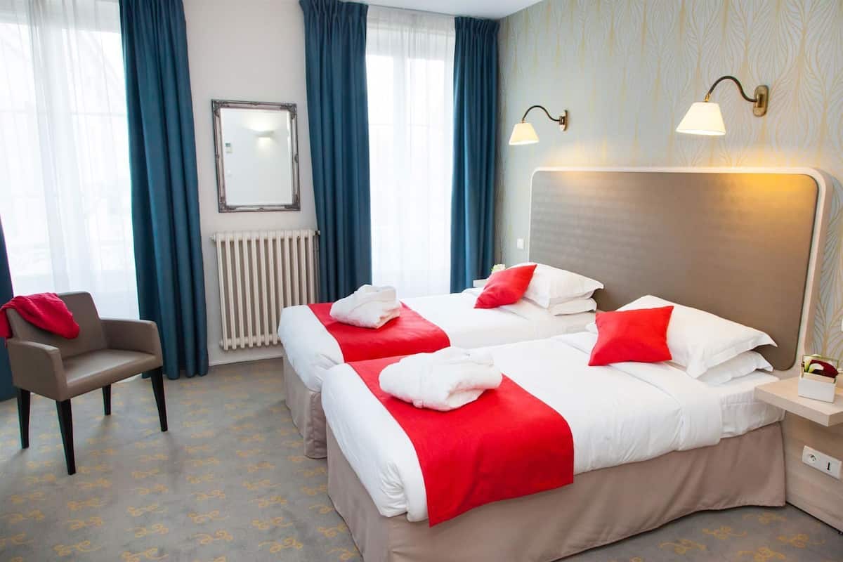 A hotel room with two beds, a chair, a radiator, and a wall-mounted lamp.
