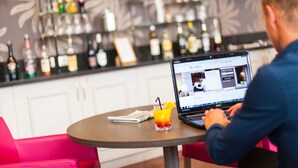 A person using a laptop at a table with a drink and a magazine. The background features a bar with various bottles.
