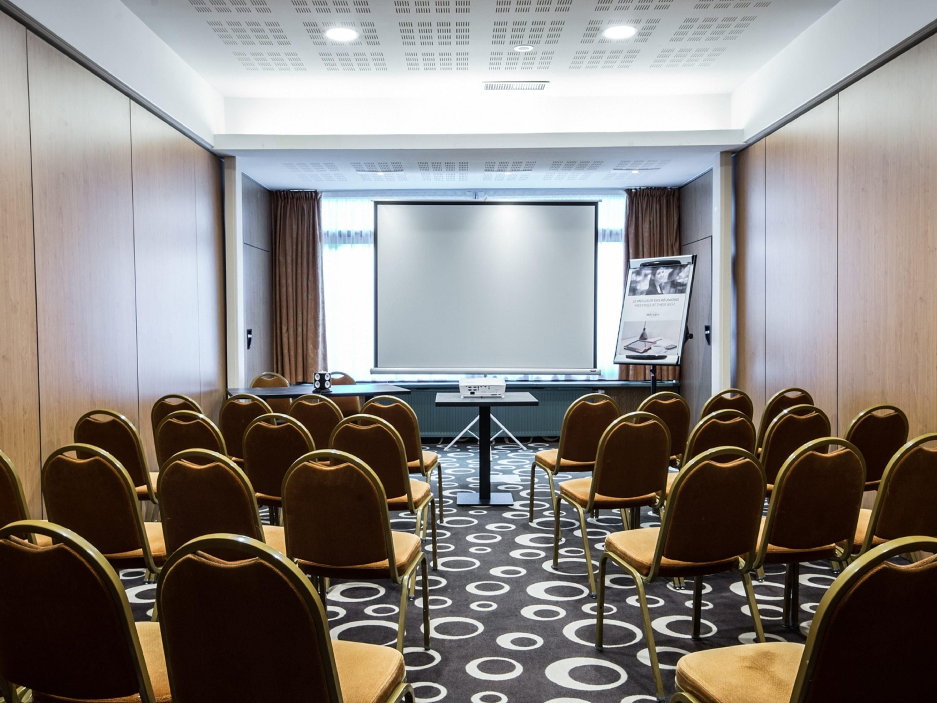 Une salle de conférence avec des rangées de chaises, un écran de projection et un tableau de présentation.