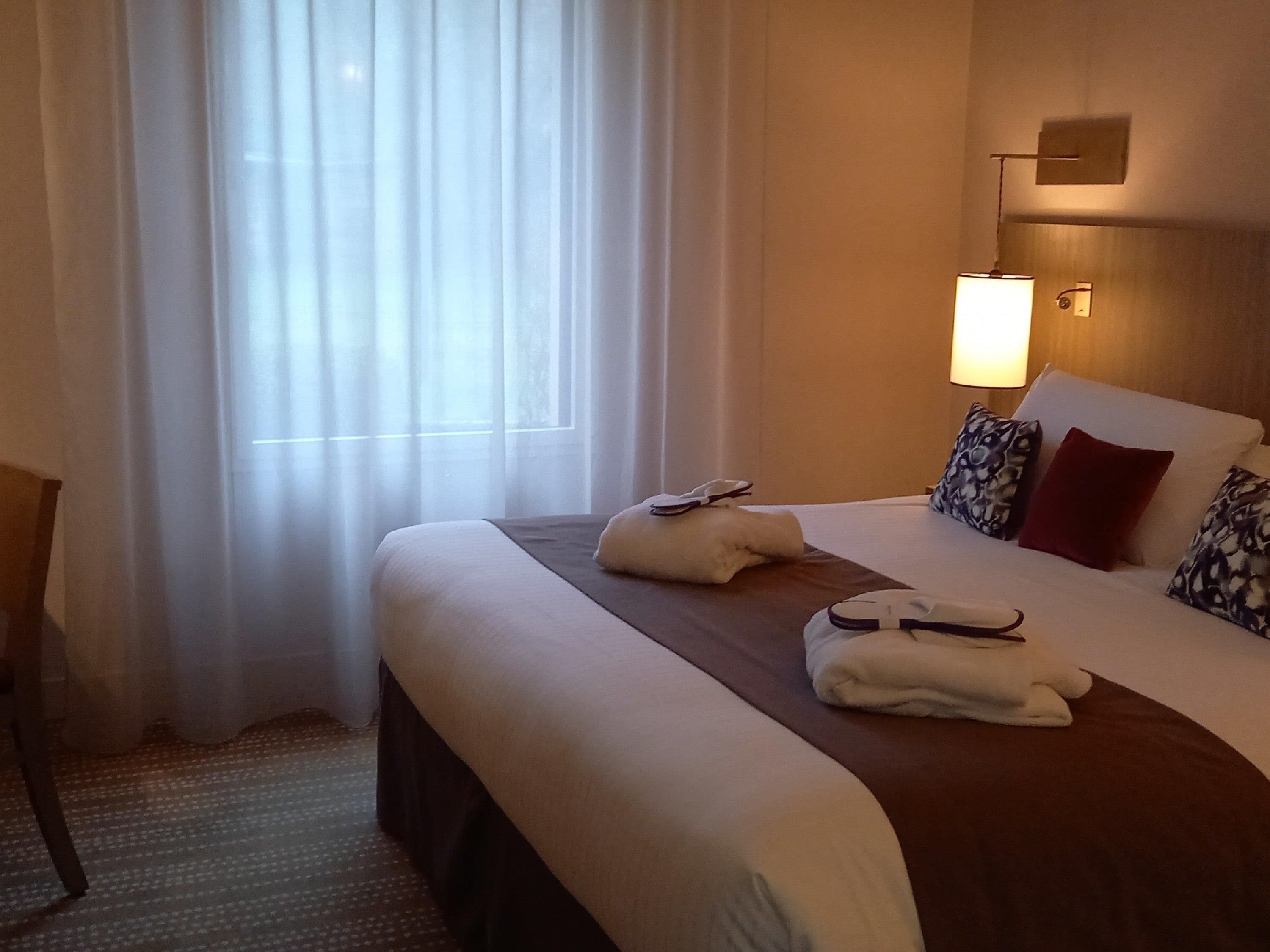 A hotel room with a large bed, a nightstand, a lamp, and sheer curtains.