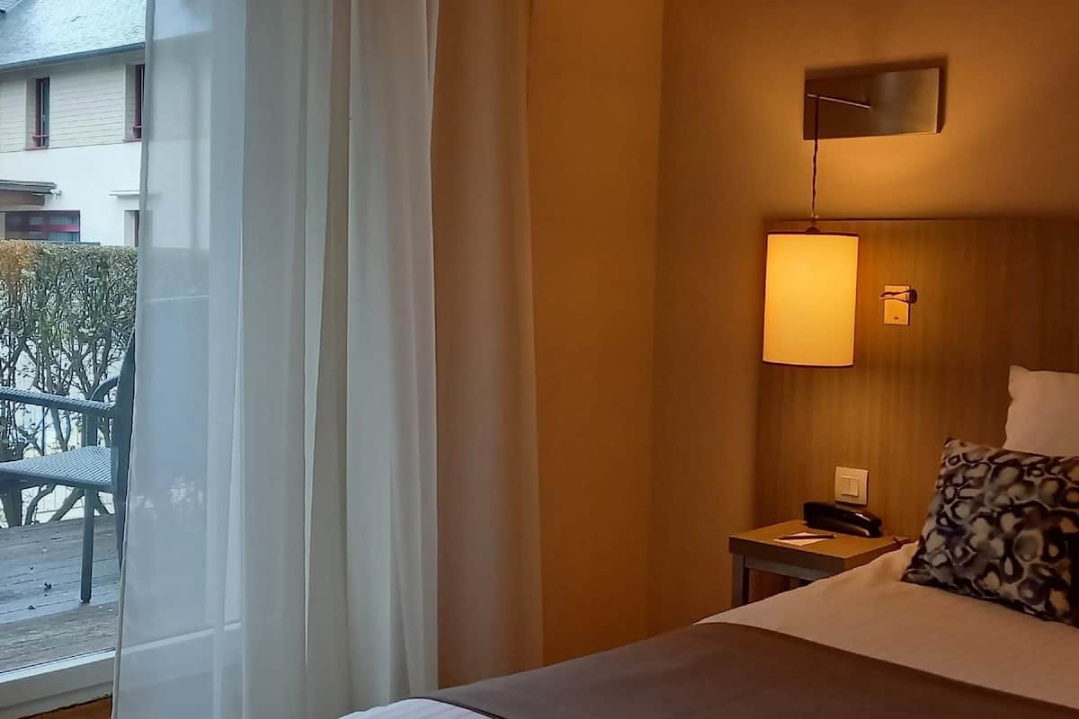 A hotel room with a bed, a nightstand, a lamp, and a view of the outdoors.