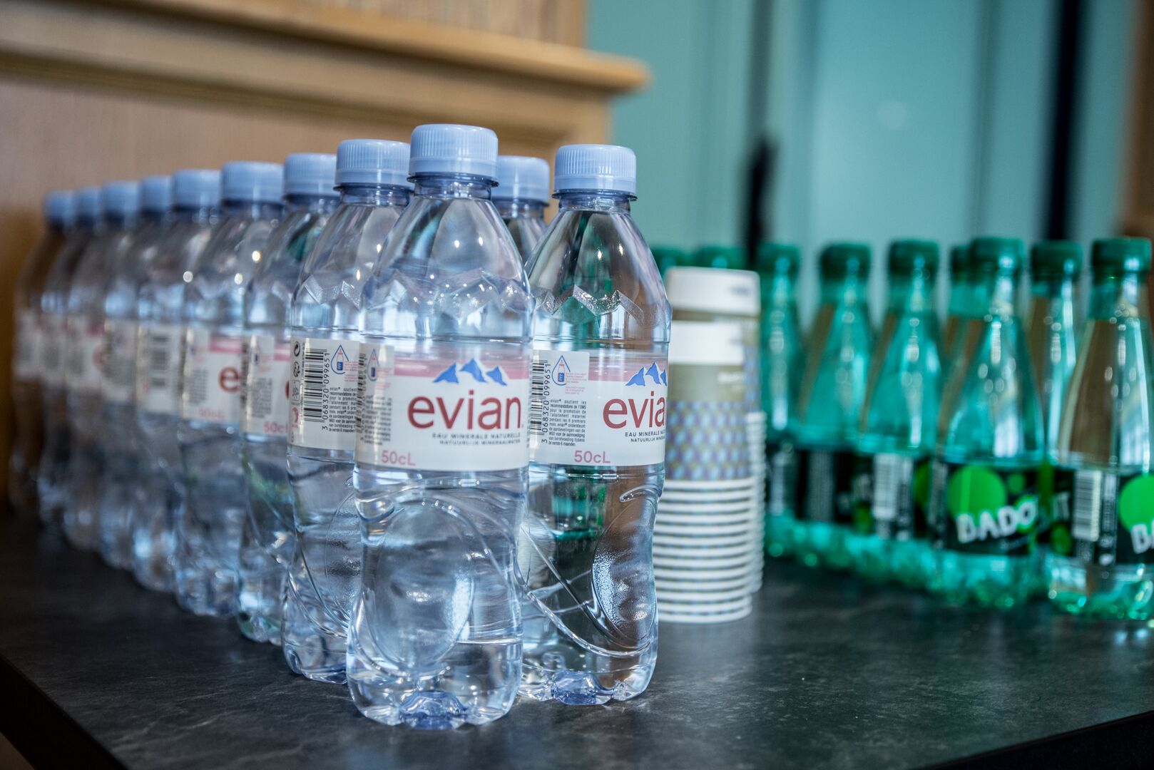 Des rangées de bouteilles d’eau Evian et de bouteilles de soda Bado sont alignées sur une table.