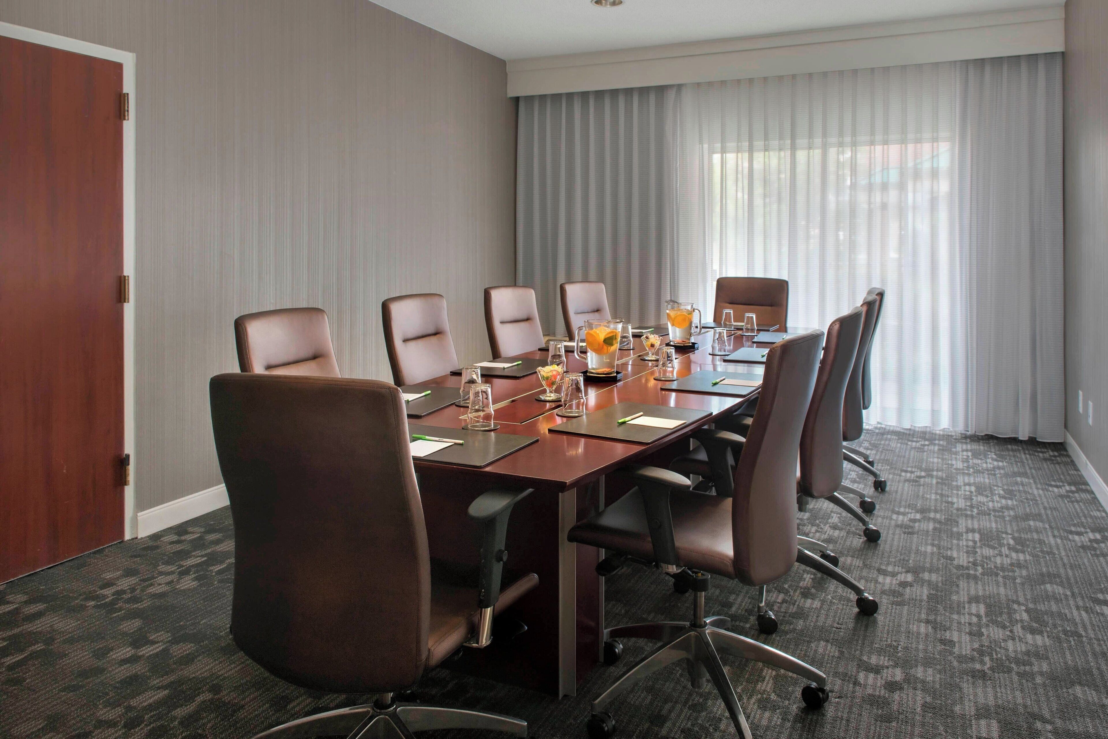 A conference room with a long table, six chairs, and a large window with curtains.