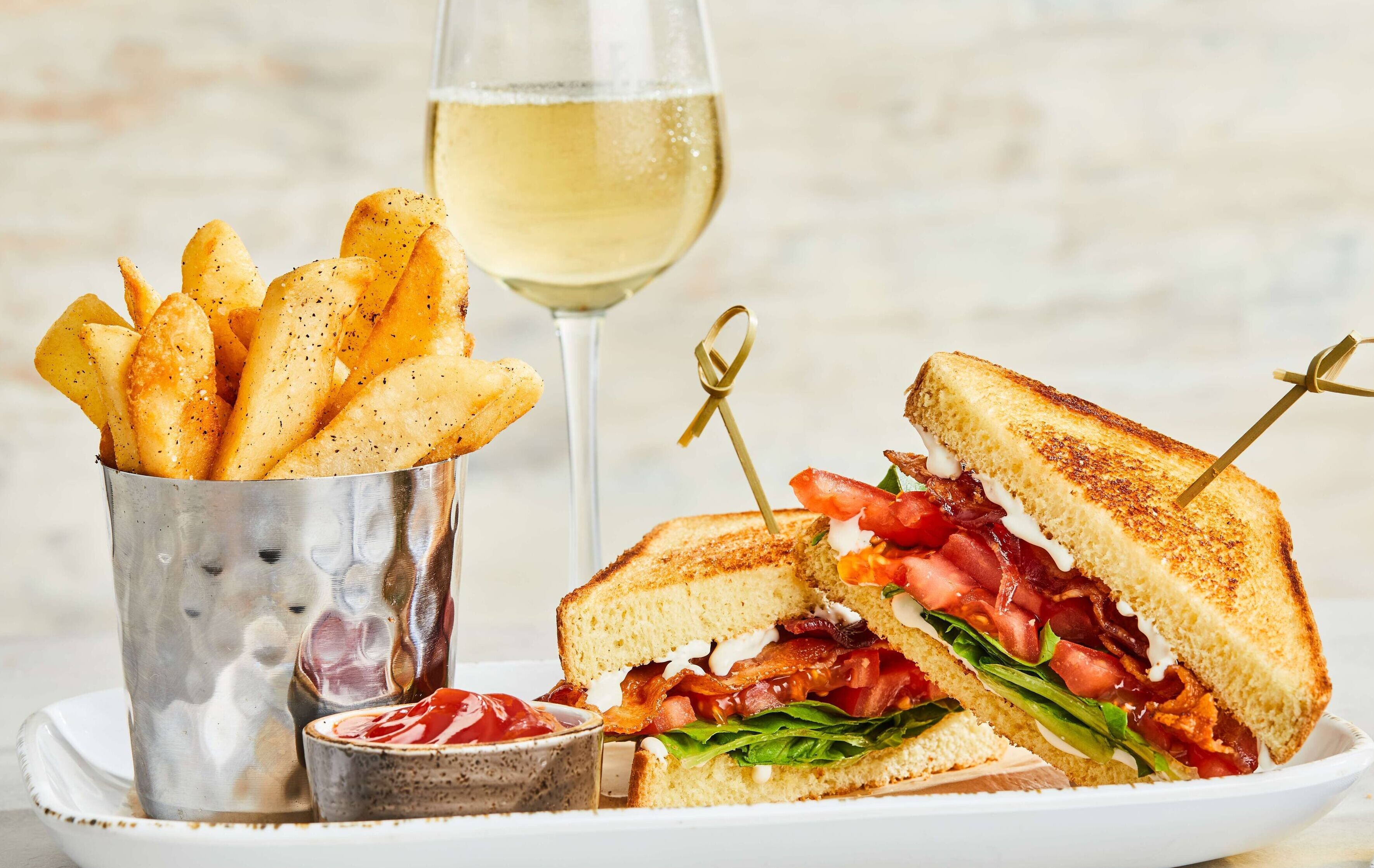 Ein Sandwich mit Zahnstocher, ein Glas Weißwein und eine Portion gewürzte Pommes.