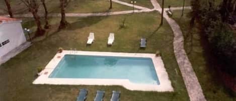 Una piscina rectangular con plataforma de azulejos, rodeada por un borde de piedra. Hay tres sillas de descanso alineadas frente a la piscina.