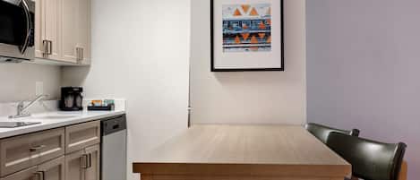 A modern kitchen with beige cabinets, a microwave, and a coffee maker. There is a framed picture on the wall and a wooden table with a chair.