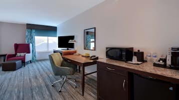 Habitación de hotel con escritorio, televisión, microondas, refrigerador, un sillón rojo, una silla verde y una ventana con cortinas.
