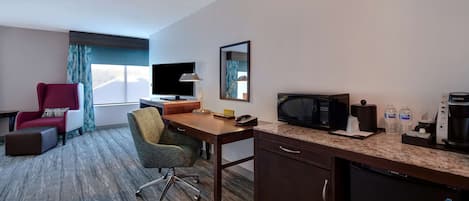 A hotel room with a desk, a television, a microwave, a refrigerator, a red armchair, a green chair, and a window with curtains.