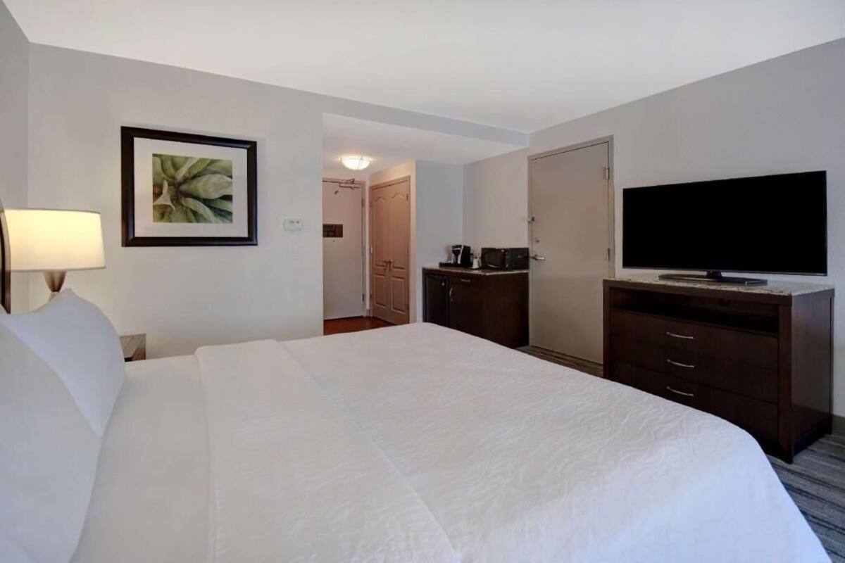 Ein Hotelzimmer mit Bett, Fernseher, Schreibtisch mit Kaffeemaschine und einem gerahmten Bild an der Wand.