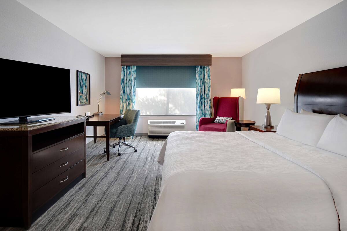 Ein Hotelzimmer mit einem großen Bett, einem Schreibtisch mit Flachbildfernseher, einem roten Sessel, einer Lampe und einem Fenster mit Vorhängen.