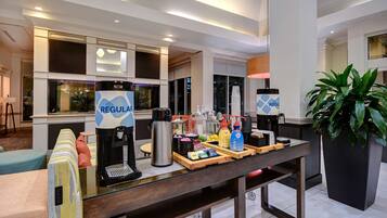 A hotel reception area with a coffee station, a reception desk, and a large potted plant.