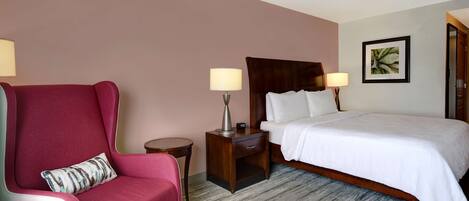 A hotel room with a bed, a red armchair, a small table with a lamp, and a framed picture on the wall.