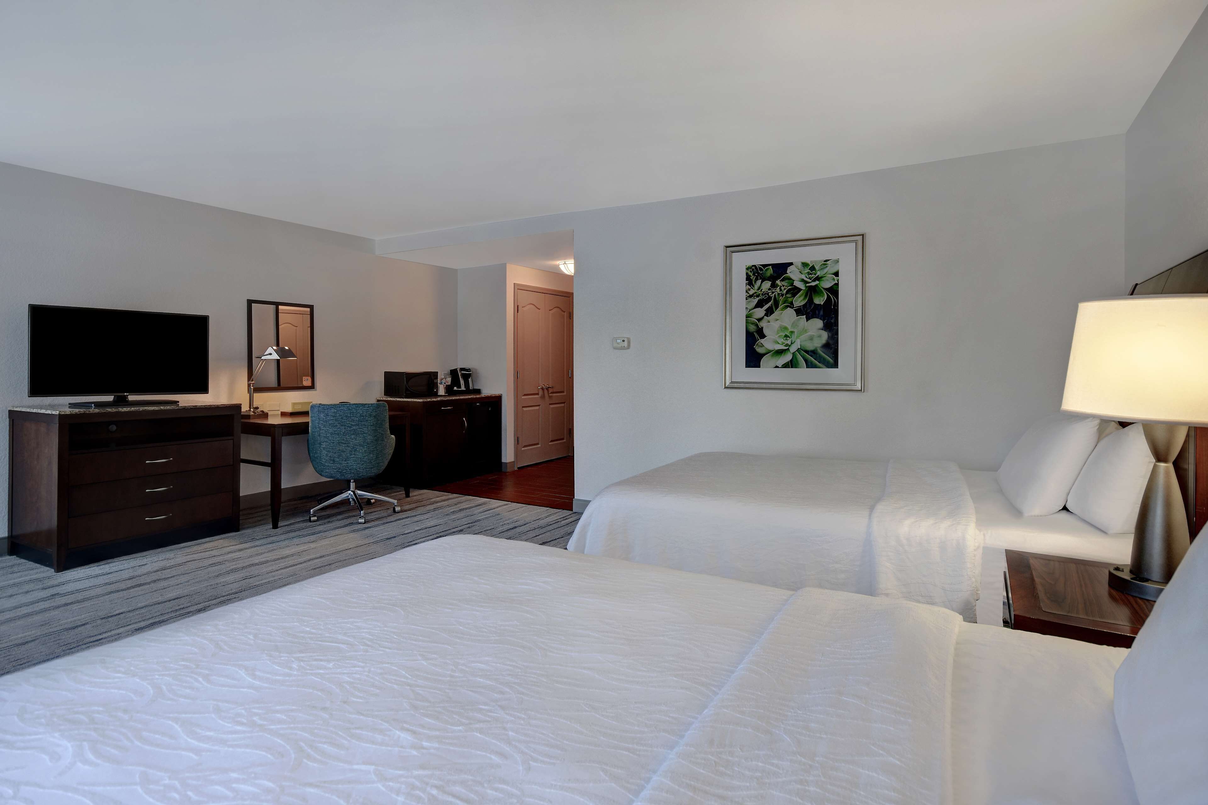 A hotel room with two beds, a desk, a chair, a TV, and a painting on the wall.