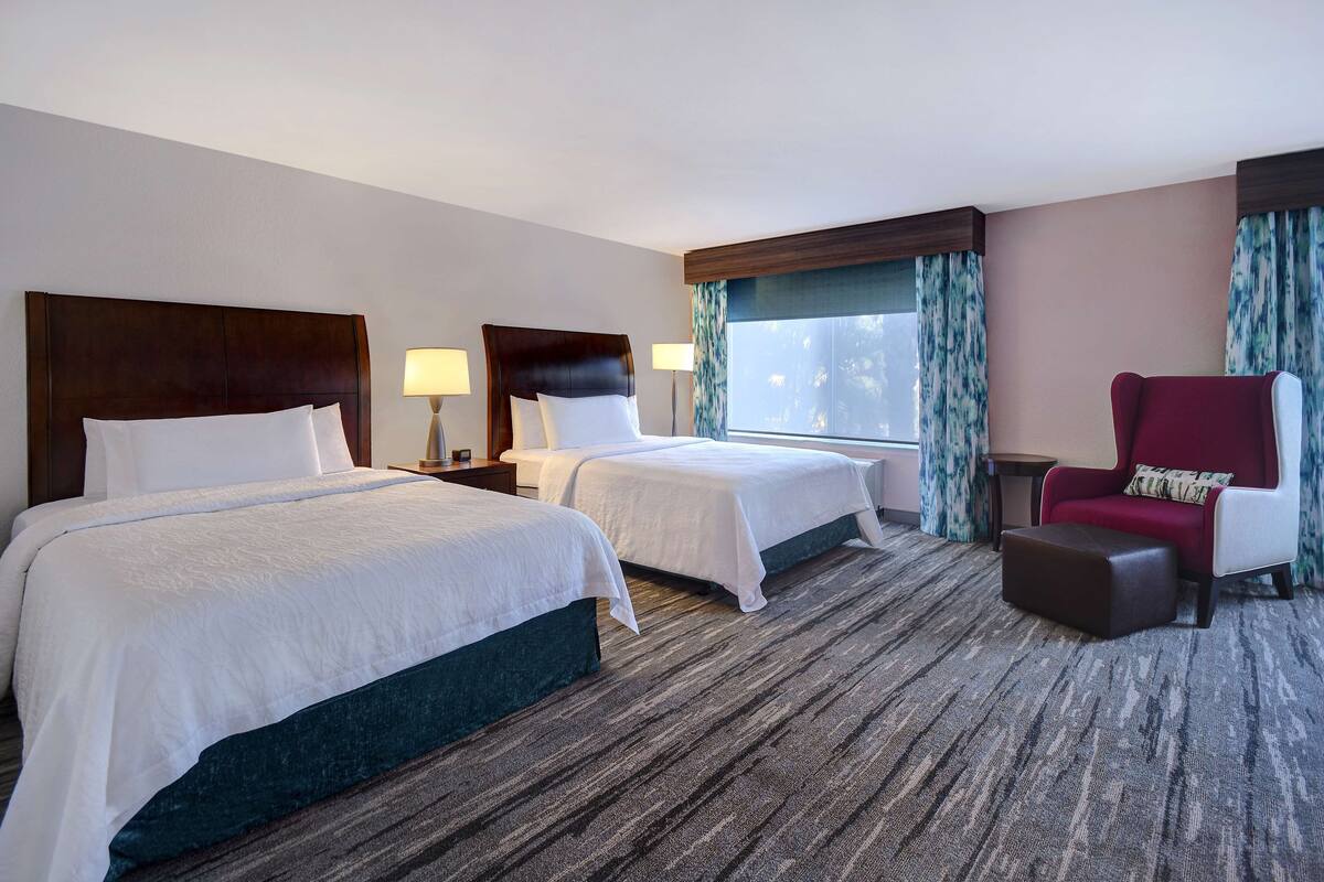 Ein Hotelzimmer mit zwei Betten, einem roten Sessel, einem kleinen Tisch und einem fenster mit Vorhängen.