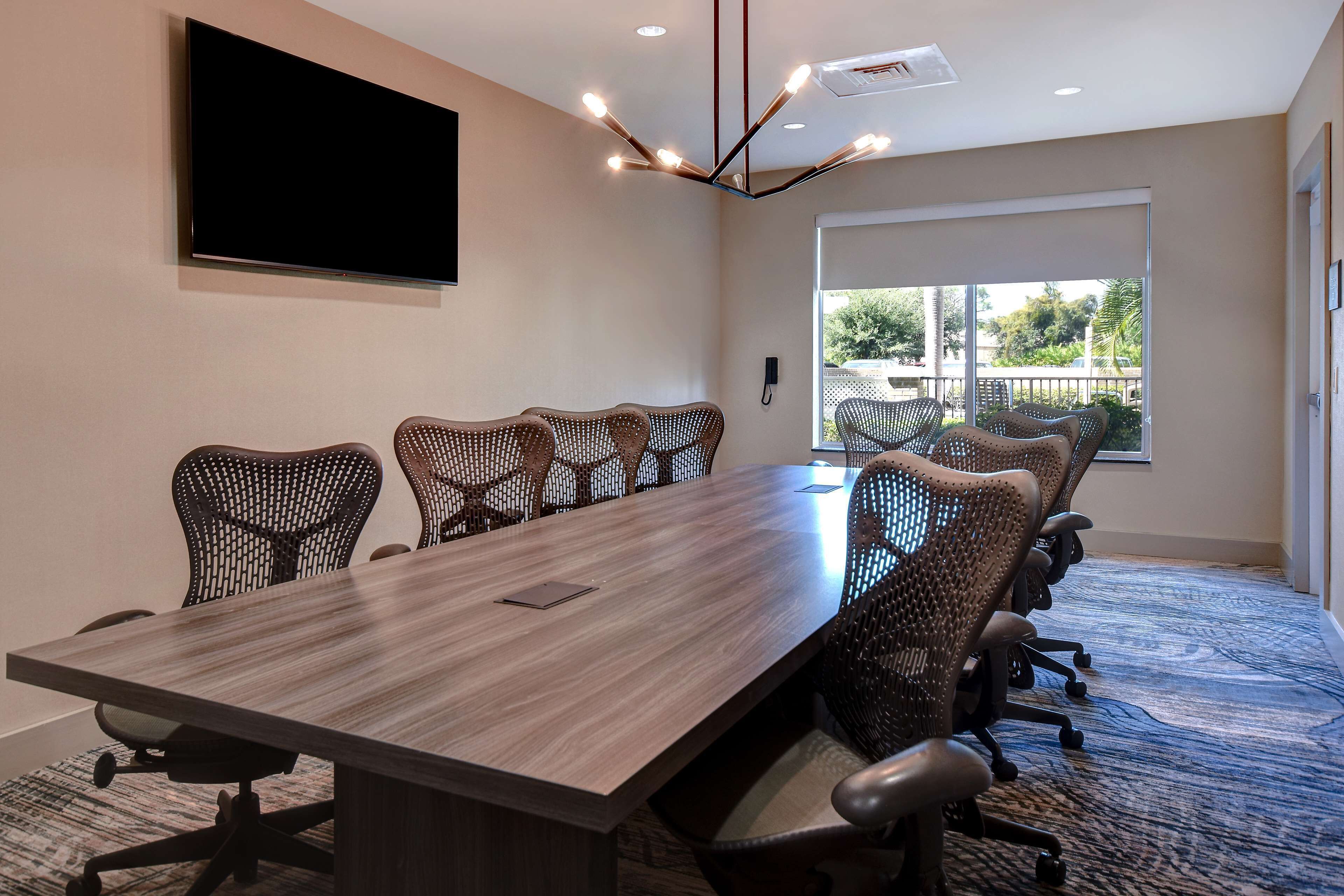 A conference room with a long table, multiple chairs, a mounted TV, and a large window with a view of greenery.