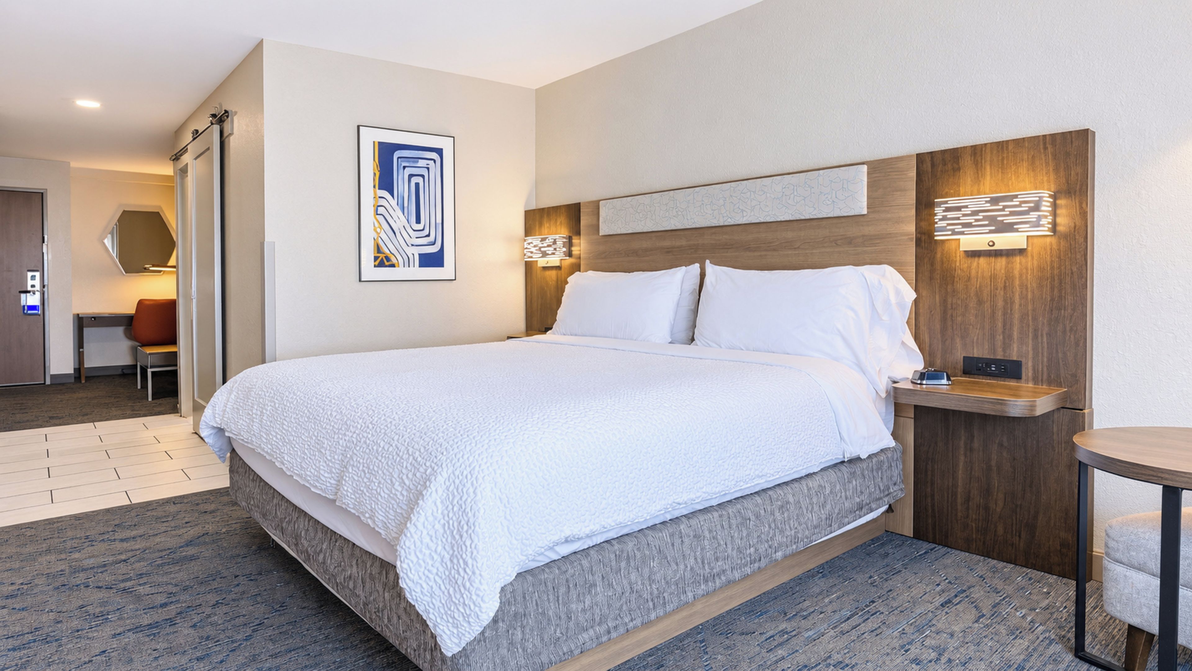 Quarto de hotel com uma cama grande, uma escrivaninha, uma cadeira e luminária fixada na parede.