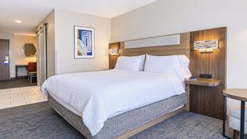 Quarto de hotel com uma cama grande, uma escrivaninha, uma cadeira e luminária fixada na parede.