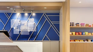 Un área de recepción de hotel moderna con un diseño geométrico en azul y blanco, un letrero visible para Holiday Inn Express Suites y una estantería con diversos alimentos envasados.