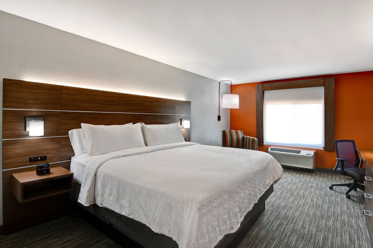 A hotel room with a large bed, a nightstand with a clock, a desk with a chair, and a window with blinds.