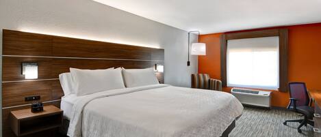 Een hotelkamer met een groot bed, een nachtkastje met een wekker, een bureau met een stoel en een raam met jaloezieën.