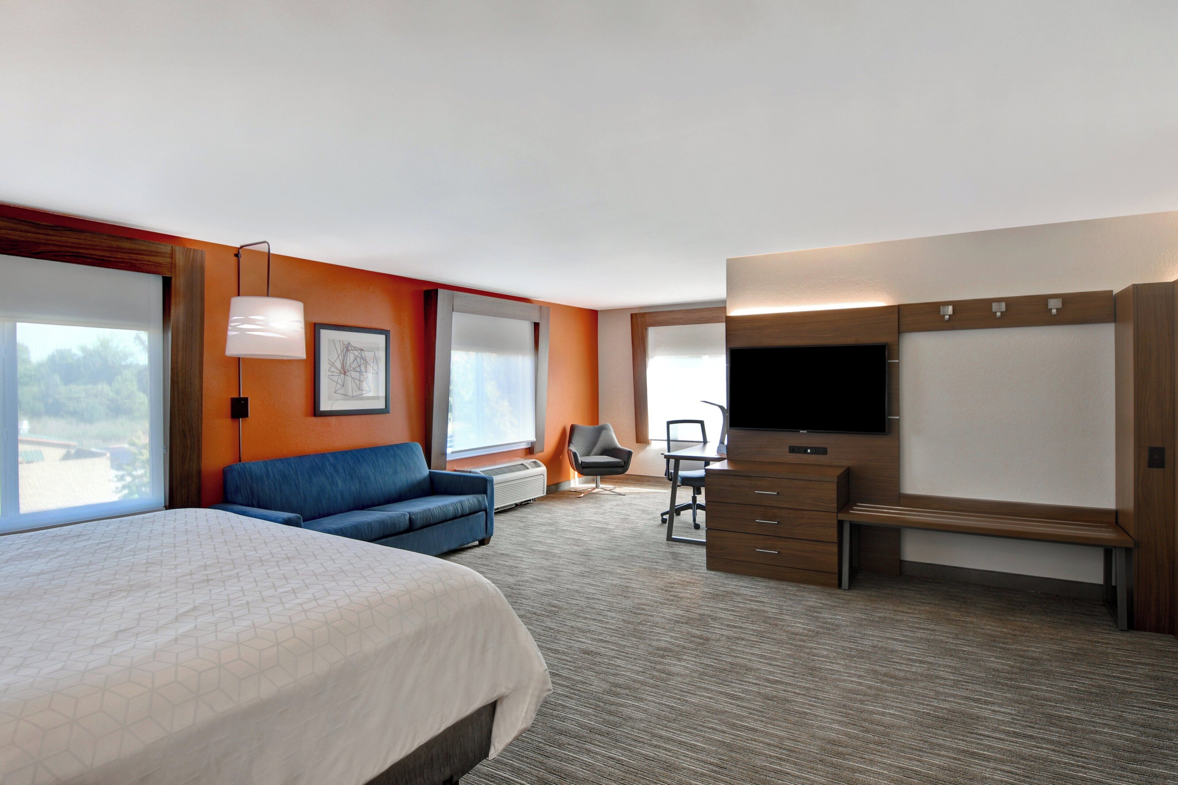 A hotel room with a bed, a sofa, a desk with a chair, a TV, and large windows.