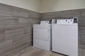 Une buanderie équipée de deux machines à laver et deux sèche-linge superposés, avec des murs recouverts de carreaux gris et un sol de couleur claire.