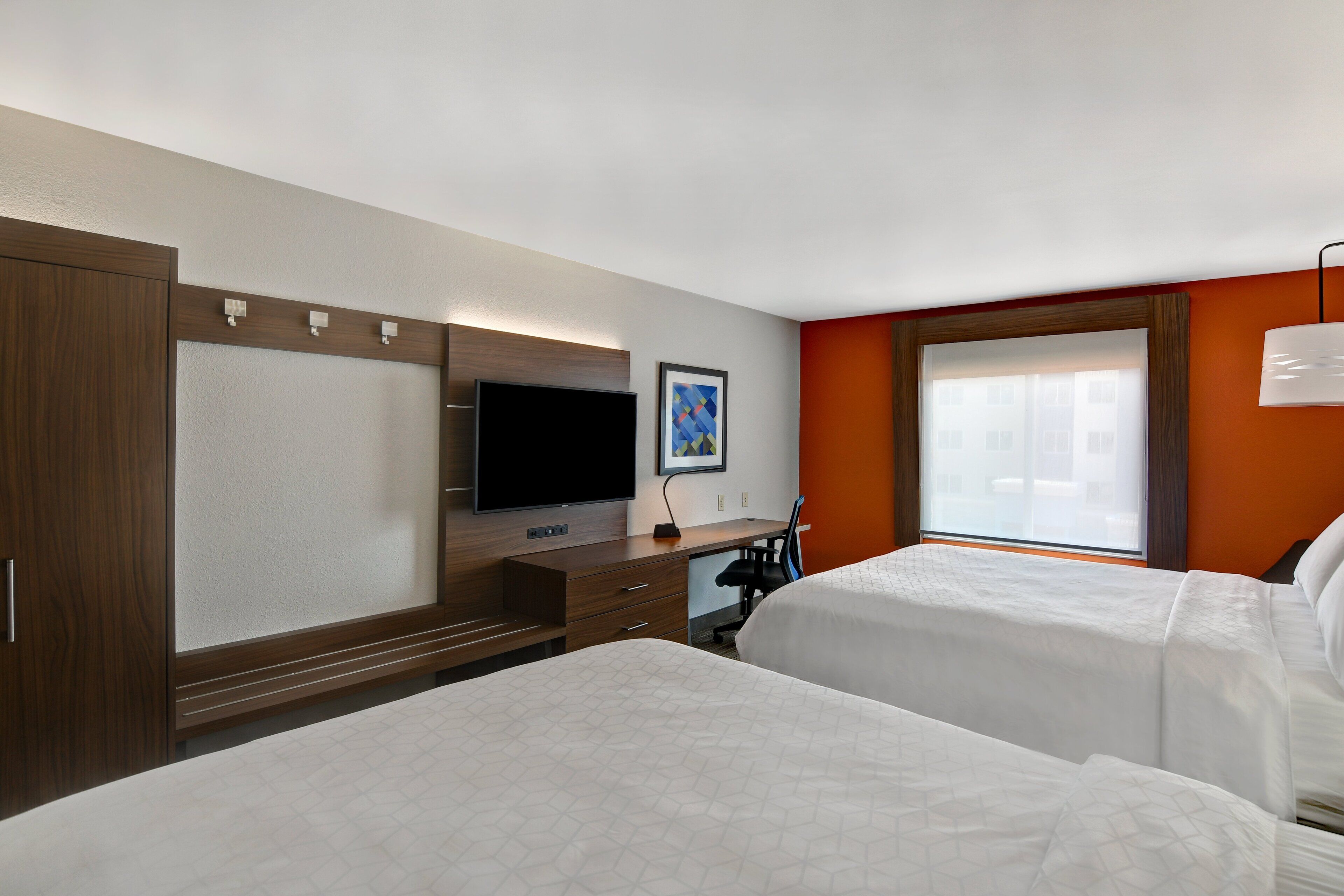 A hotel room with two beds, a desk, a TV, and a window with a view of buildings.