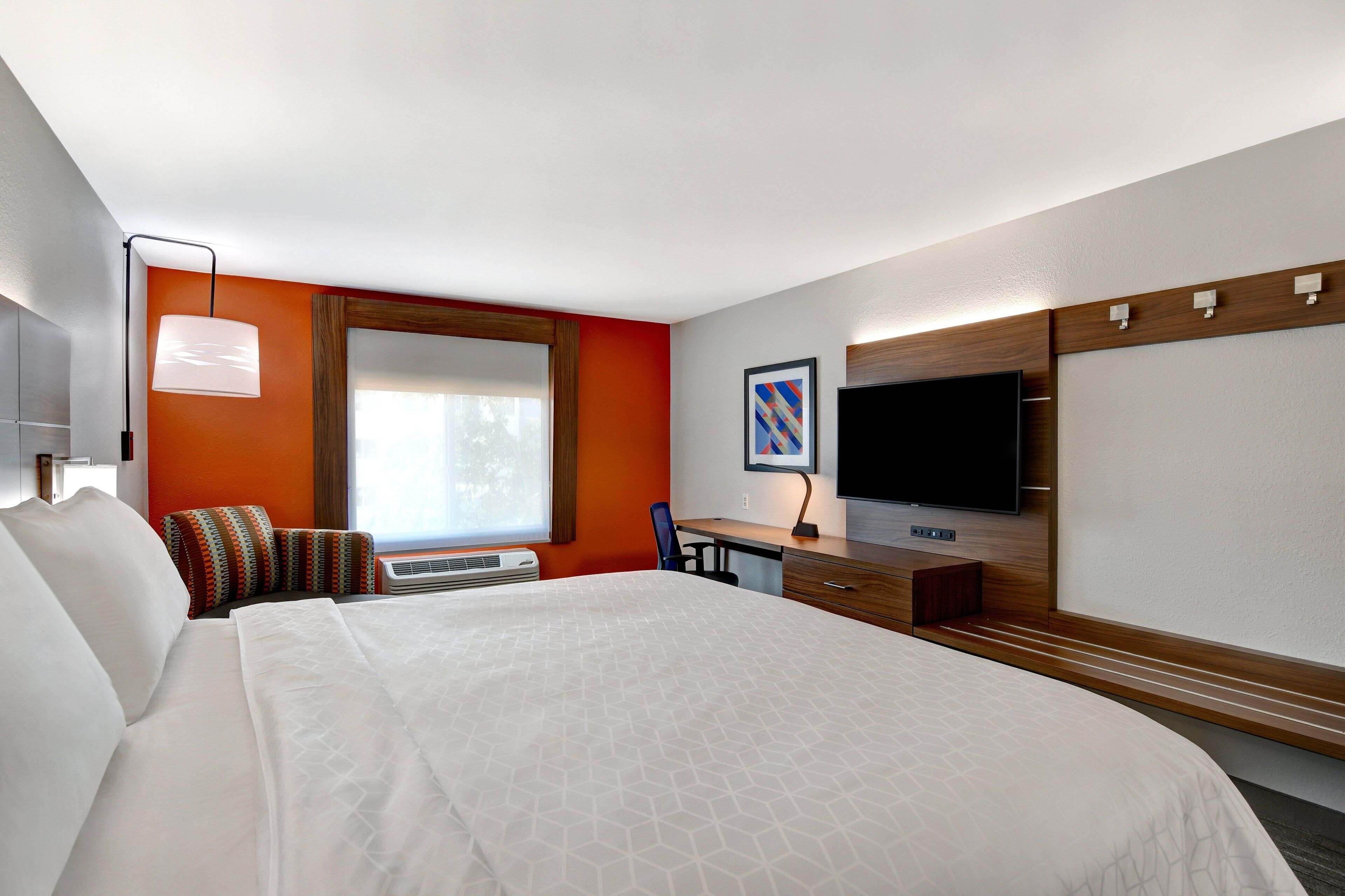 Ein Hotelzimmer mit einem großen Bett, einem Schreibtisch, einem Flachbildfernseher und einem Fenster mit Jalousien.