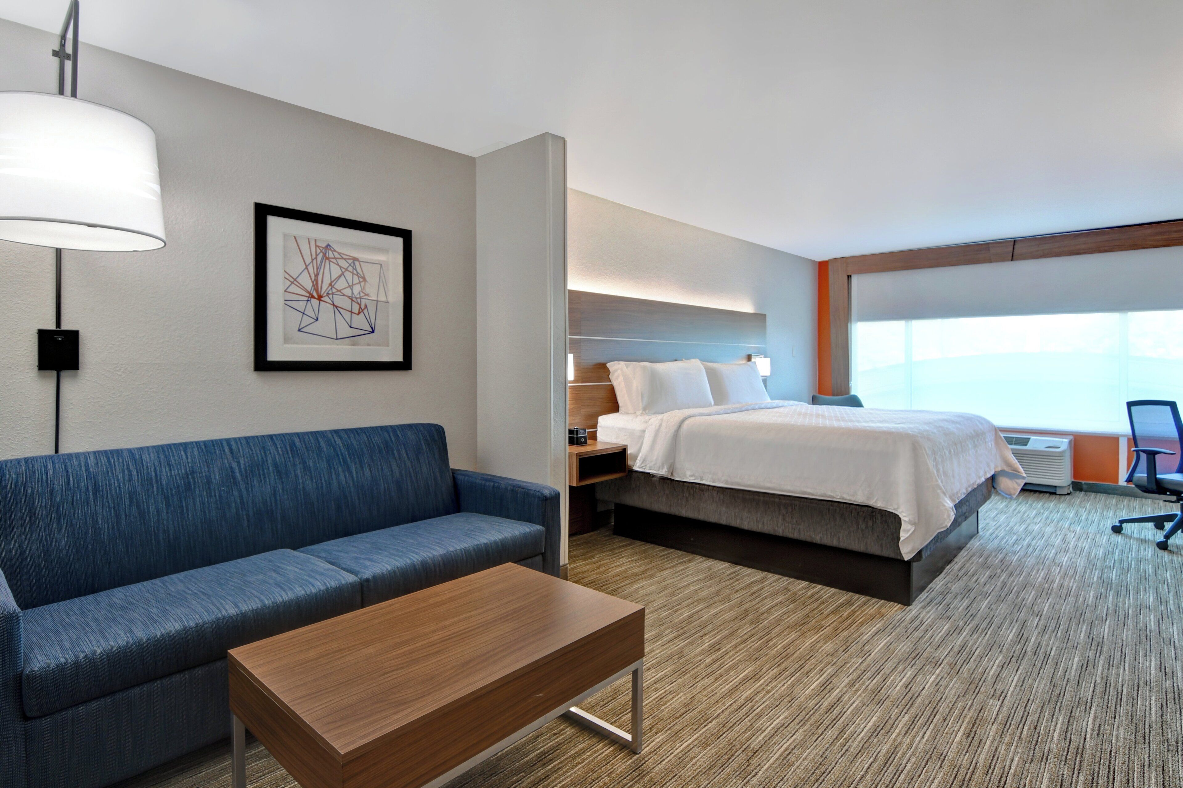 A hotel room with a blue sofa, a wooden table, a bed with white bedding, a framed abstract artwork on the wall, and a large window providing a view.