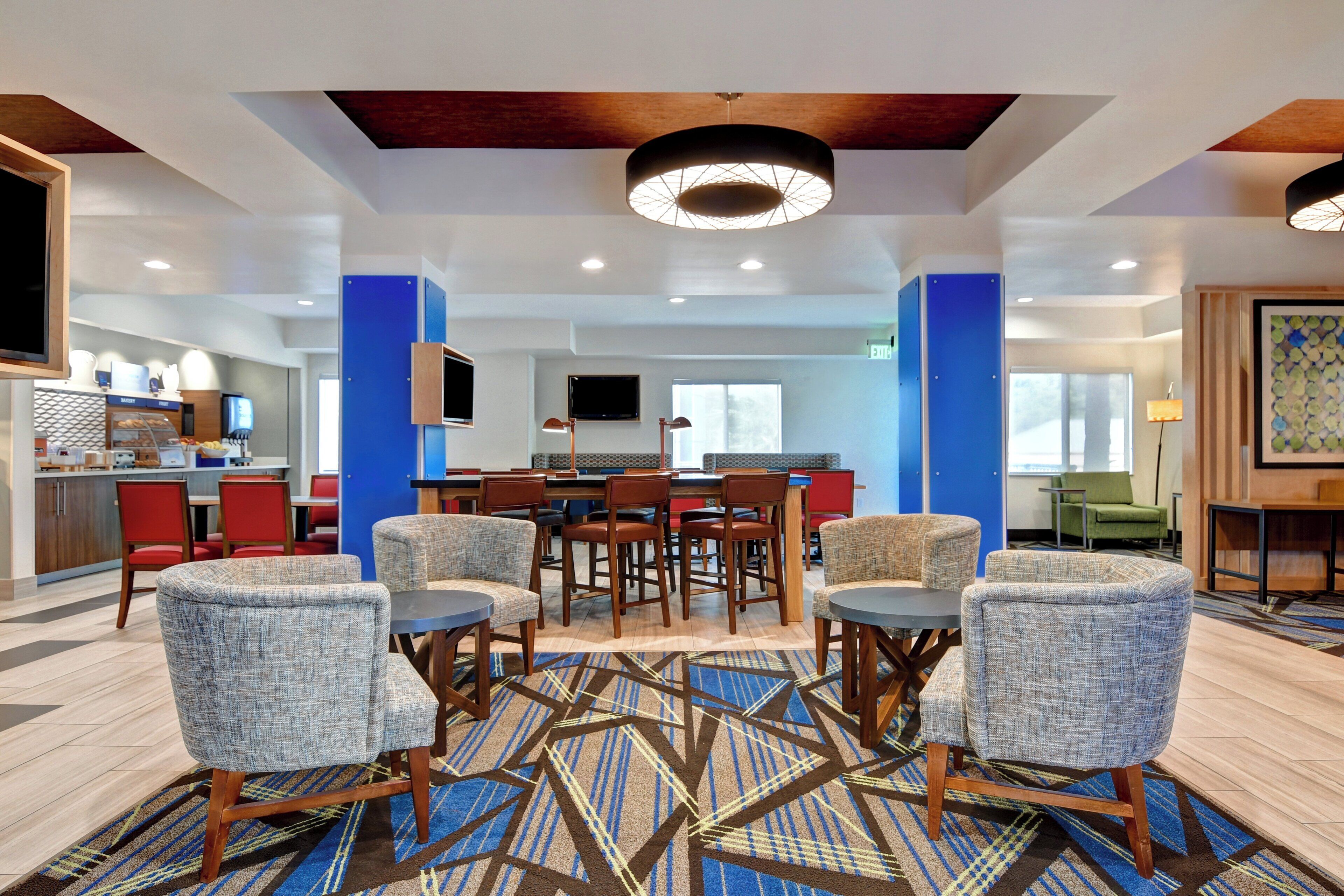 Un hall de hotel con un área de comedor central, diversas opciones de asientos y una cocina al fondo.