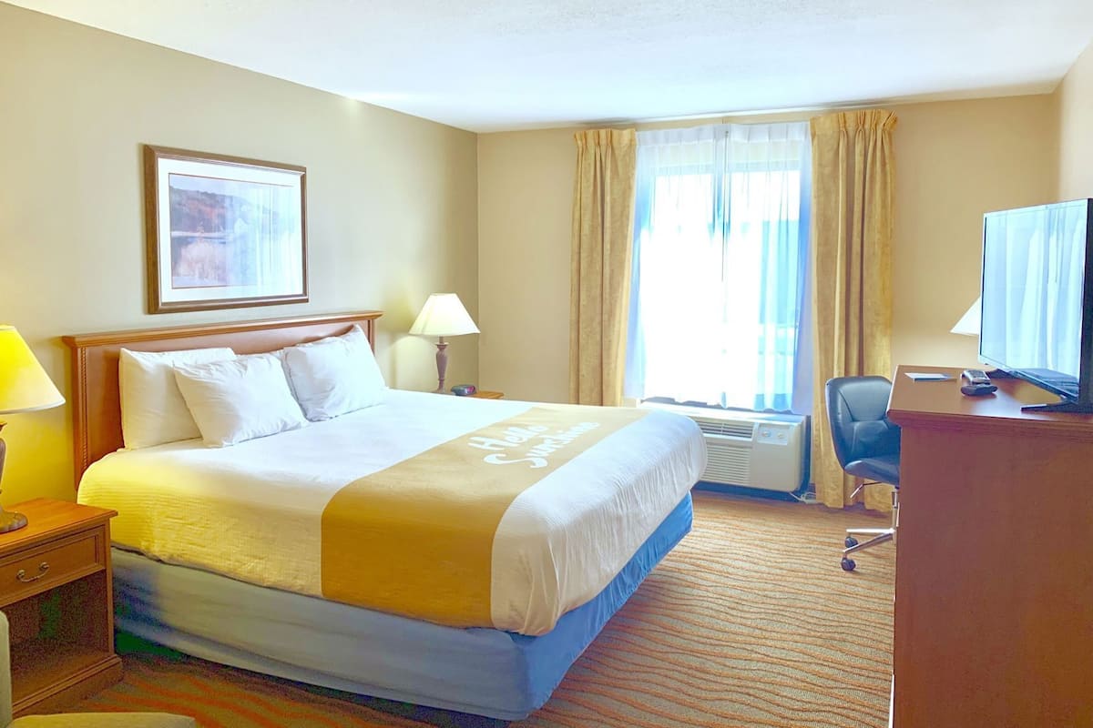 Ein Hotelzimmer mit einem Bett, einem Schreibtisch mit Fernseher, einem Sessel und einem Fenster mit Vorhängen.