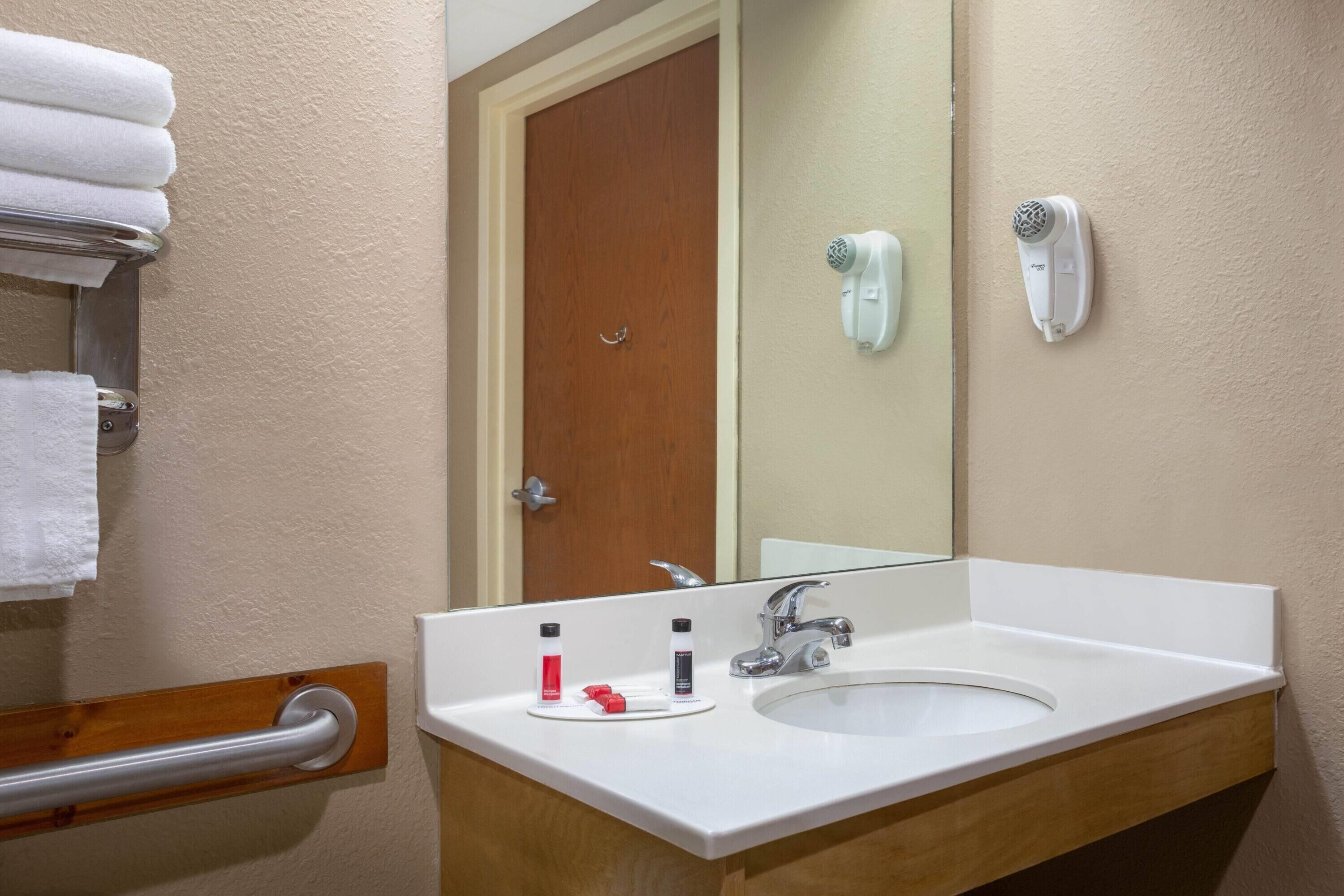 Un baño con un lavabo blanco, un espejo, un perchero con toallas blancas, un secamanos y una puerta.