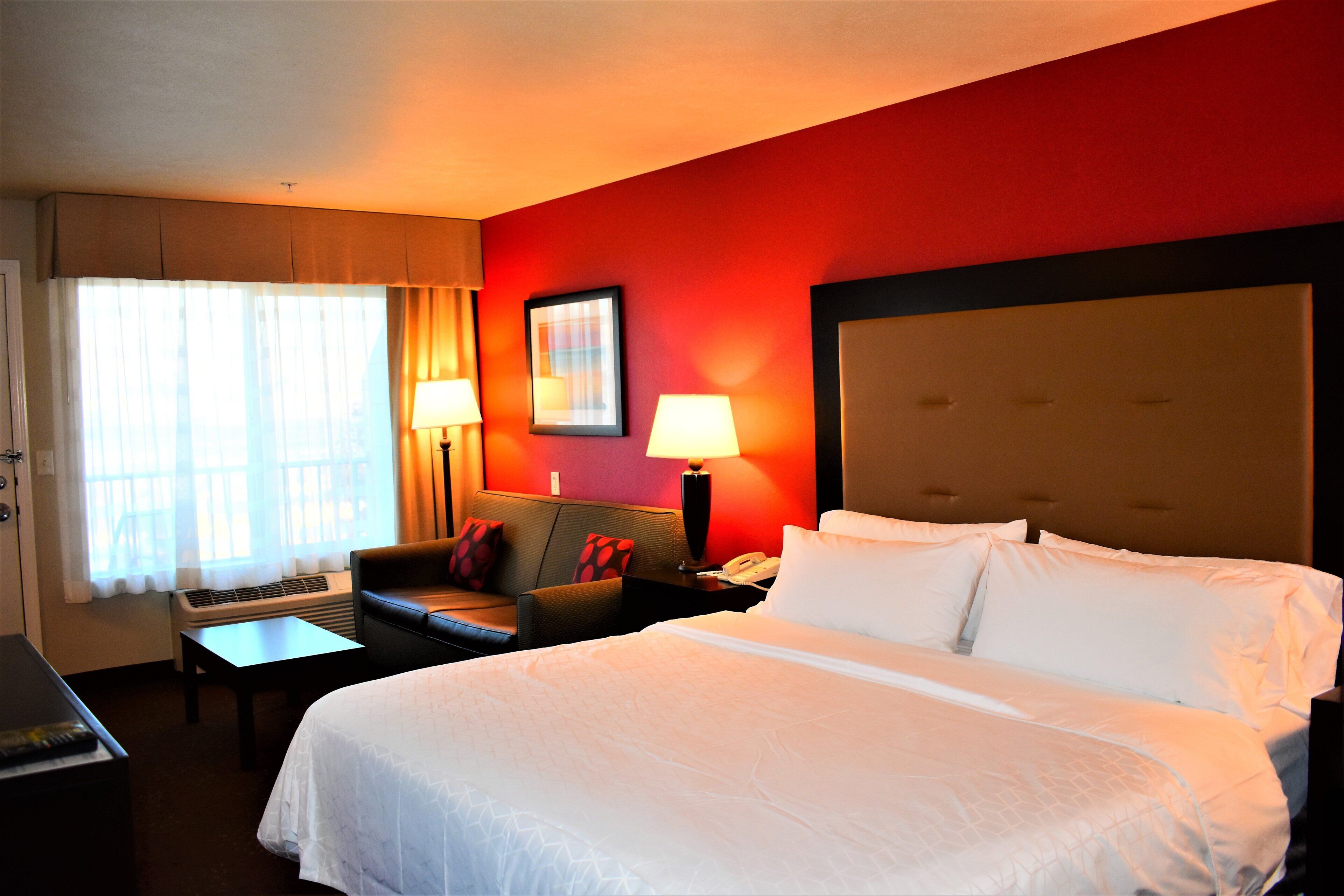 A hotel room with a large bed, a desk, a chair, a nightstand, a lamp, and a window with curtains.