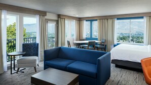 A hotel room with a bed, a blue sofa, a small table, a chair, and a balcony with a view of the city and mountains.