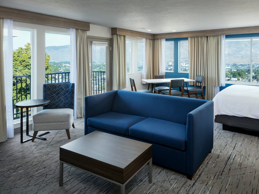 A hotel room with a bed, a blue sofa, a small table, a chair, and a balcony with a view of the city and mountains.