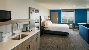 A modern hotel room with a kitchenette, a bed, a desk, and a view of the outdoors.