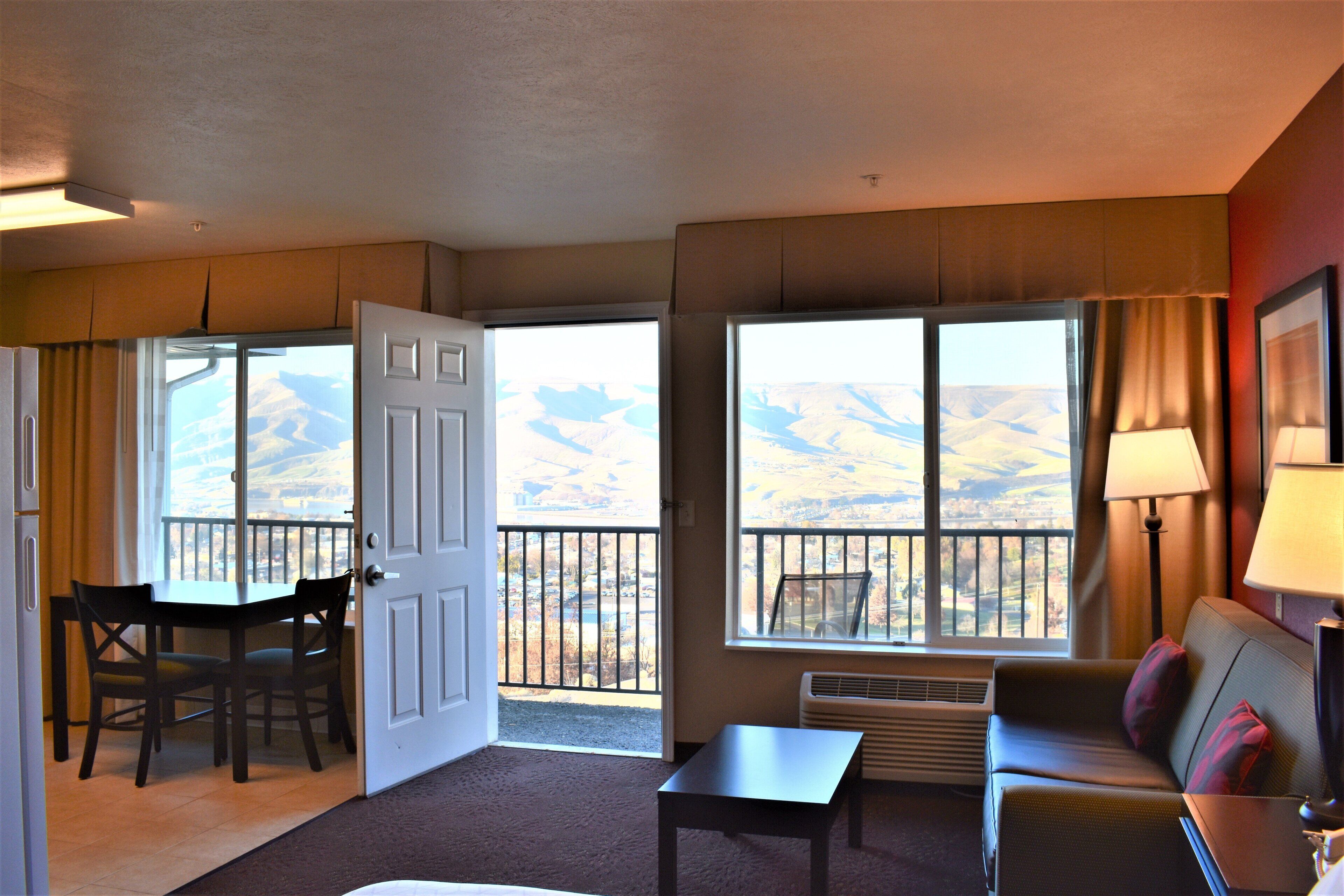 A hotel room with a dining table, chairs, a sofa, a standing lamp, and a view of a mountainous landscape through large windows.