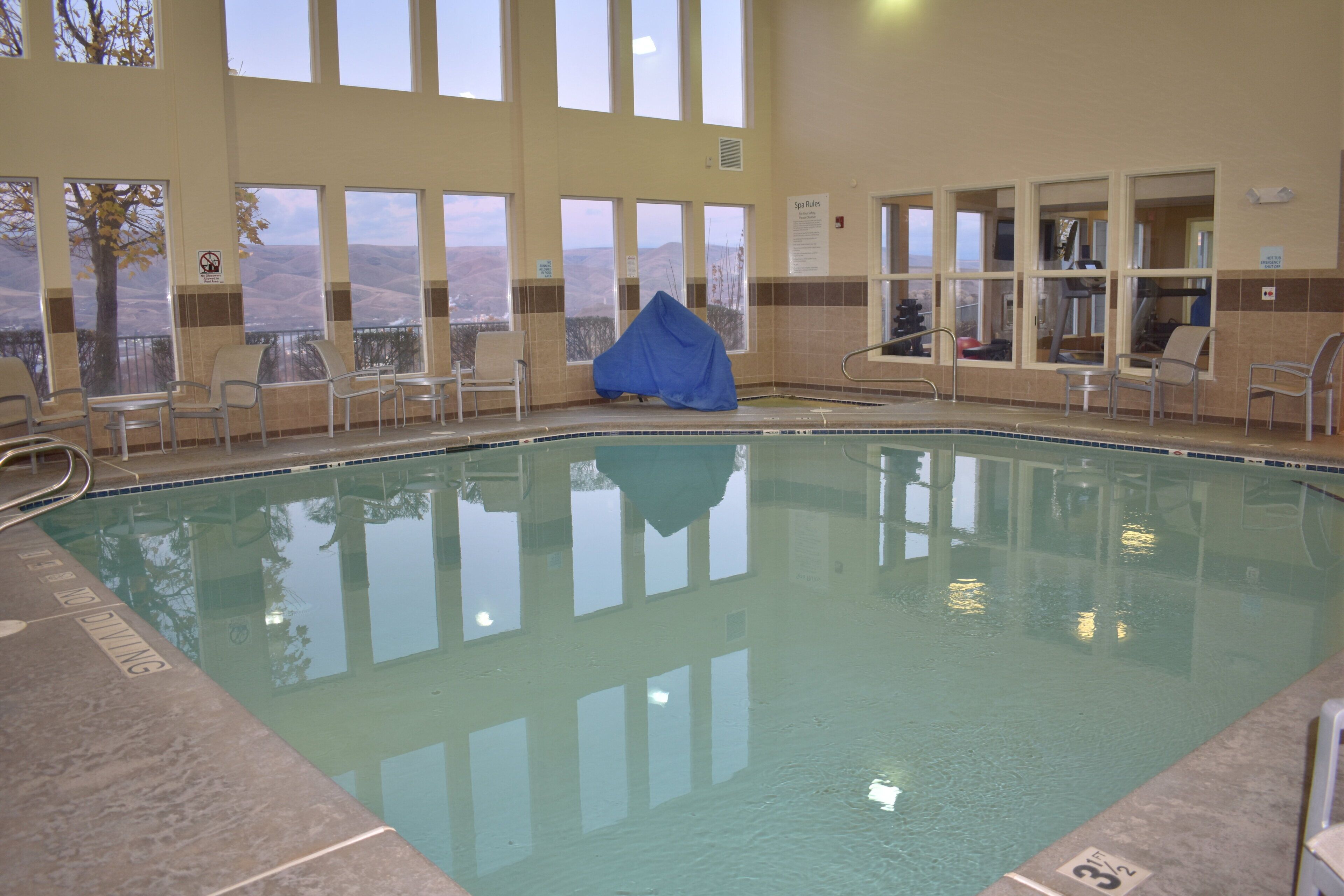 En indendørs swimmingpool med store vinduer, stole og et overdækket område.