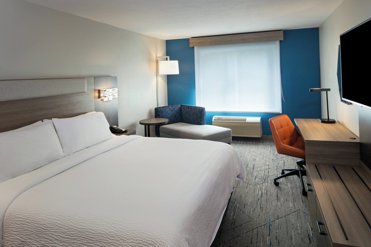 Une chambre d’hôtel avec un grand lit, un bureau, une chaise orange, une petite table ronde et une grande fenêtre avec des stores.