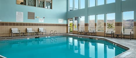 Une piscine intérieure à l’eau d’un bleu cristallin, entourée de murs de couleur beige et bleue, et offrant une vue sur un paysage désertique grâce à de grandes fenêtres.