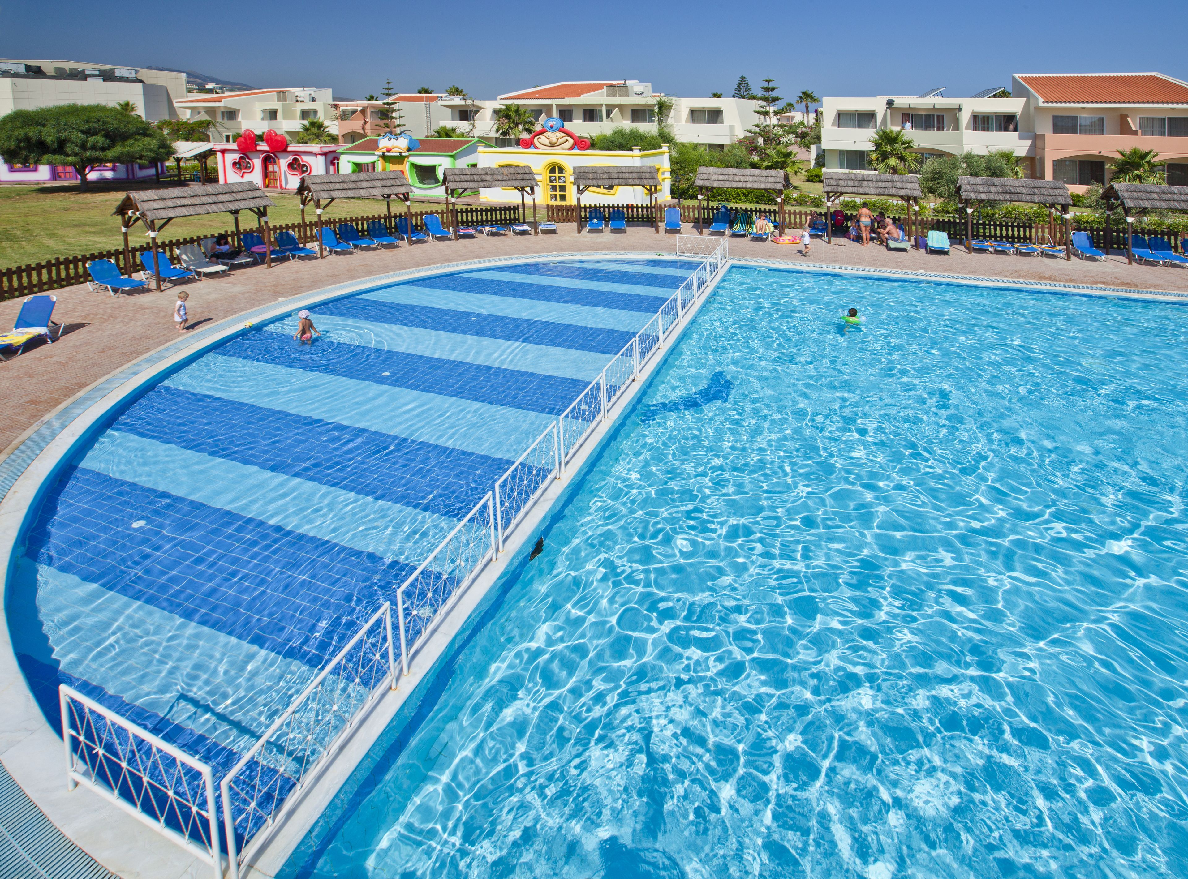 Una grande piscina trasparente con bordo in piastrelle blu e bianche, circondata da lettini e ombrelloni.