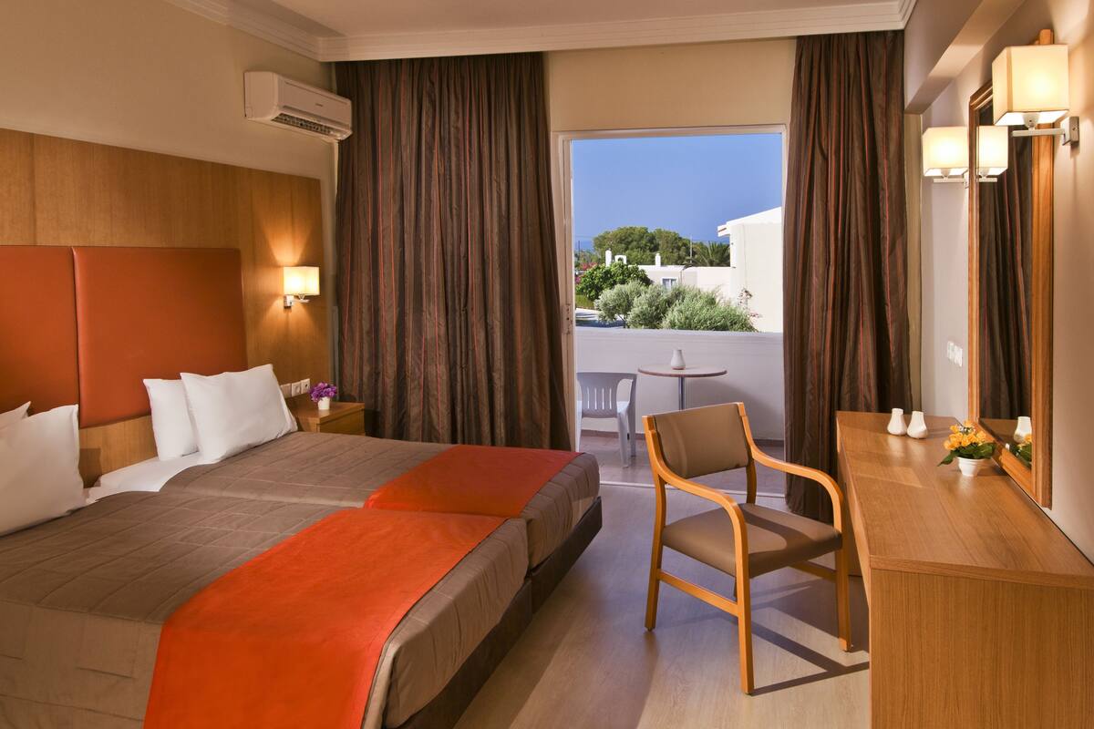 A hotel room with a large bed, a desk, a chair, and a balcony with a view of buildings and trees.