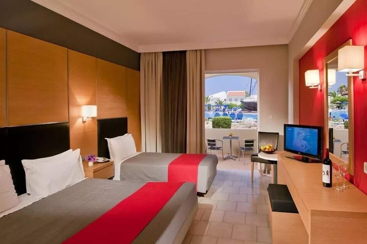 Une chambre d’hôtel avec deux lits, une télévision, une petite table avec une bouteille et un verre, et une vue sur l’espace piscine.