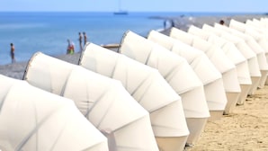 De nombreux parasols blancs sont alignés sur une plage de sable, offrant une vue dégagée sur la mer et les montagnes lointaines.