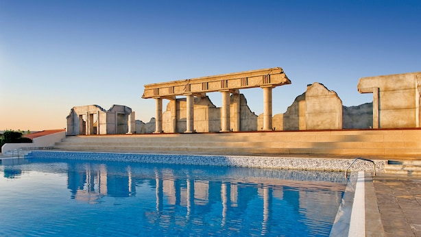 Une piscine à l’eau d’un bleu cristallin, entourée de ruines de pierre ornées de colonnes et d’arcs.