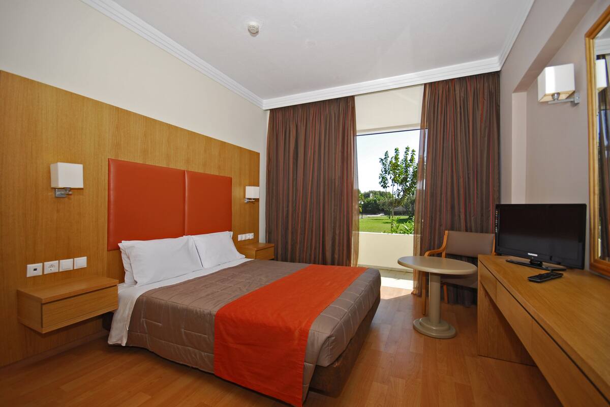 A hotel room with a bed, a desk with a TV, a chair, and a window with curtains.