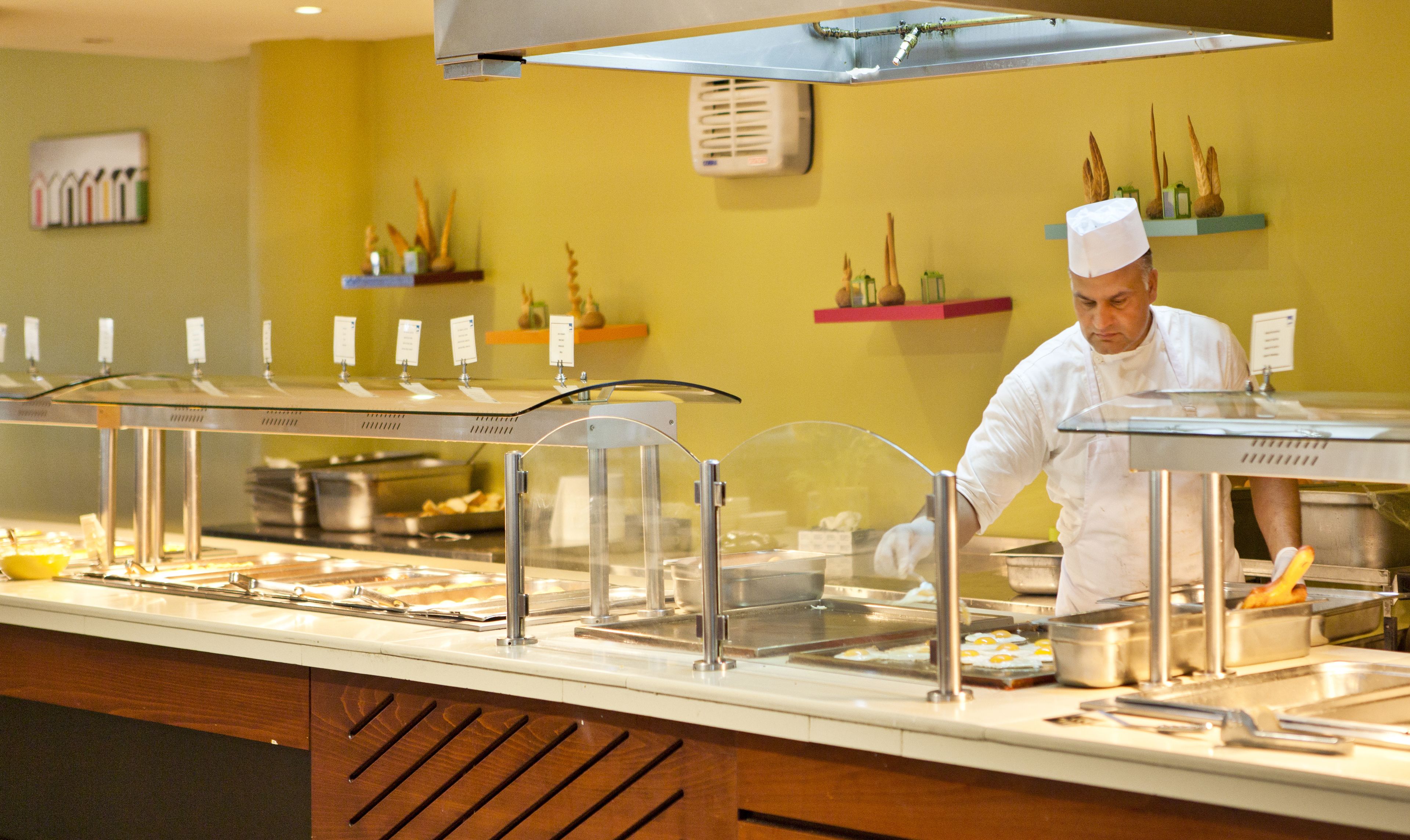Buffet montado com diversas estações de comida, um chef preparando alimentos e uma parede amarela com objetos decorativos.