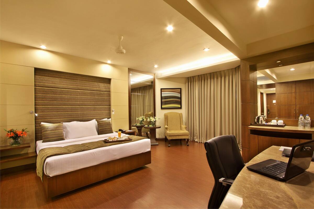 A hotel room with a large bed, a desk with a laptop, a chair, a small table with a vase of flowers, and a mirror.